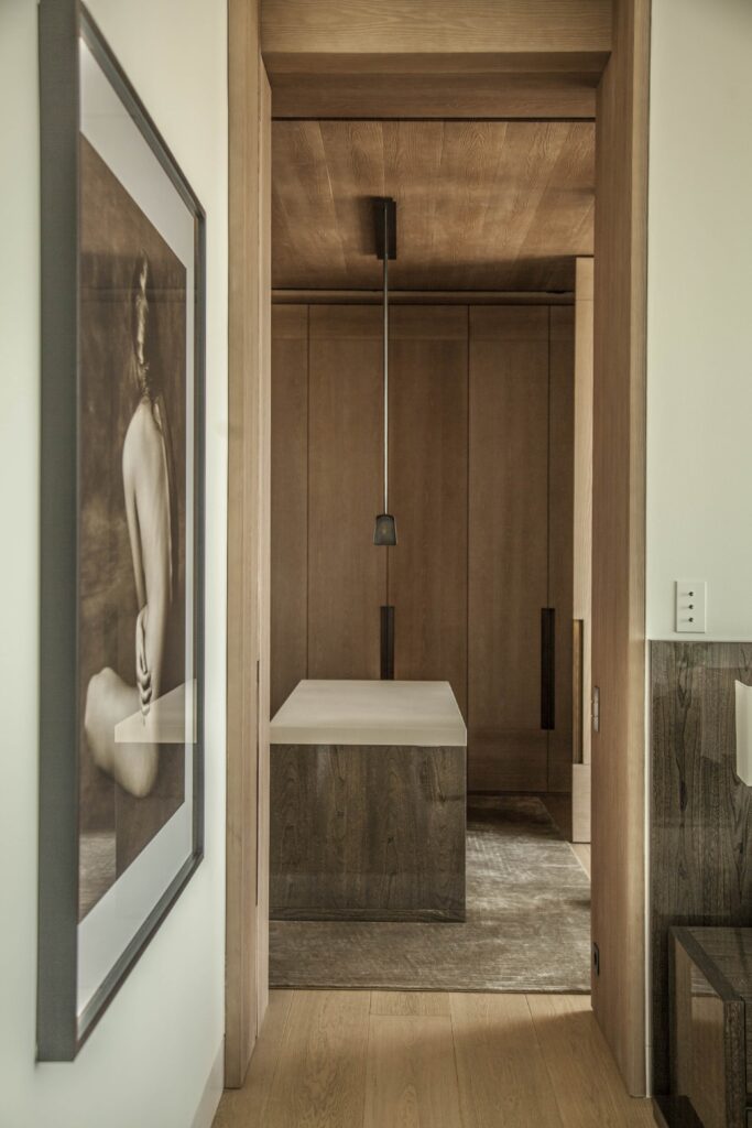 Framed by a doorway, the walk-in dressing room of the Warsaw Cosmopolitan Penthouse is a study in tonal harmony. Floor-to-ceiling bleached oak cabinetry, carried seamlessly across walls and ceiling, creates a cocoon-like atmosphere of warm, enveloping calm. At its centre, a freestanding island unit combines a dark-stained oak base with a pale stone top, anchored by a slender matte black pendant light that draws the eye downward. A silvery, worn-texture rug softens the wide-plank oak floor beneath. In the foreground, a large-format black-and-white photographic artwork introduces an intimate, human counterpoint to the architecture's geometric precision — a quiet reminder that this space, for all its restraint, is designed to be lived in.