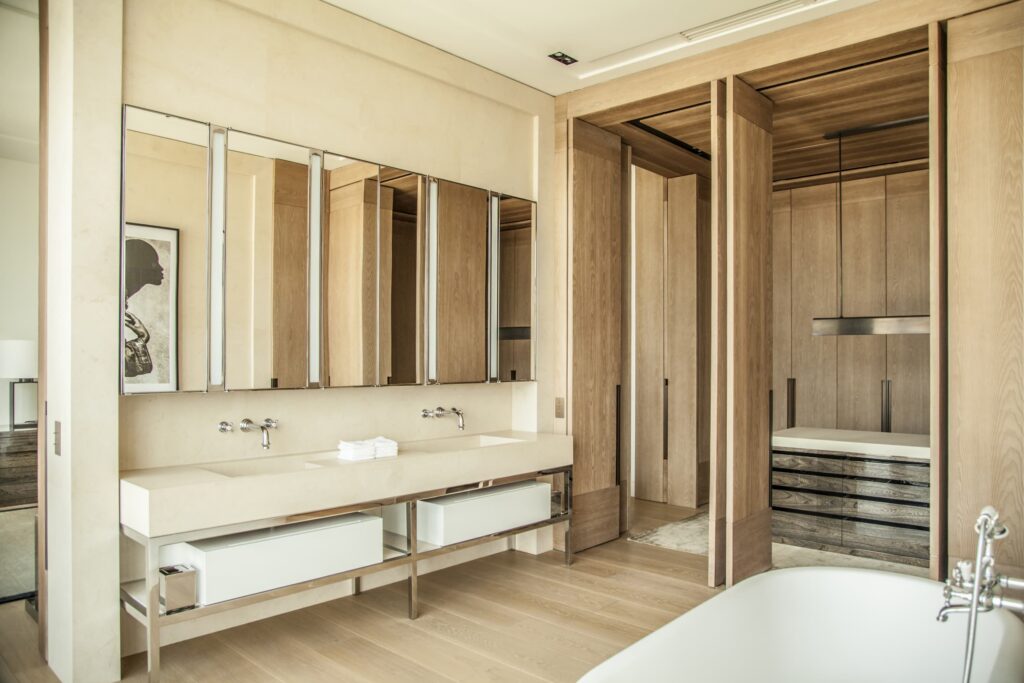 The master bathroom of the Warsaw Cosmopolitan Penthouse embodies a refined dialogue between stone and wood. A double floating vanity in honed limestone, paired with wall-mounted polished chrome fixtures, anchors the space with quiet elegance. A triptych of oversized mirror panels amplifies the natural light and reflects the warm tones of the bleached oak panelling that lines every surface. Beyond the open partition, the walk-in dressing room reveals bespoke cabinetry with dark-stained drawer units, crowned by a slender pendant light — a deliberate counterpoint to the room's otherwise pale, serene palette. A freestanding soaking tub, glimpsed in the foreground, completes this intimate retreat where every material choice speaks to understated luxury.