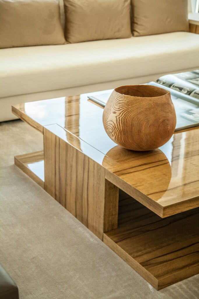 A close study of the bespoke coffee table at the heart of the Warsaw Cosmopolitan Penthouse living room. Crafted from richly grained solid wood — its warm amber tones deepened by a high-gloss lacquered surface — the table is composed of staggered, stepped tiers that give it an almost sculptural presence. Inset glass panels introduce a lightness that offsets the weight of the timber. Resting at its centre, a hand-turned wooden bowl, its concentric grain rings exposed in full, reads as both decorative object and quiet homage to the natural material language of the space. A coffee table book and a cream linen sofa in the background complete the composed, editorial stillness of the frame.